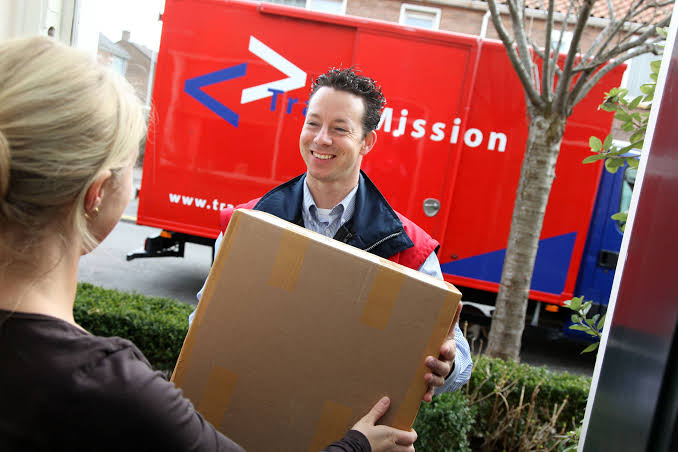 TransMission-bezorger die een pakket overhandigt bij een voordeur, met de rode TransMission-vrachtwagen op de achtergrond